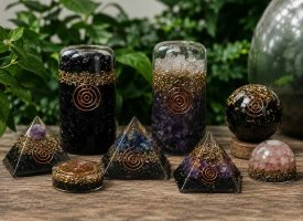 Forskellige h�ndlavede orgoniter i pyramider, kugler og t�rne med krystaller og kobberspiraler p� rustikt tr�bord med gr�nne planter i baggrunden.