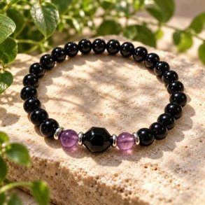 Krystalarmb�nd med sorte onyx-perler og lilla ametyst liggende p� en lys, sandfarvet stenoverflade med gr�nne planter i baggrunden.