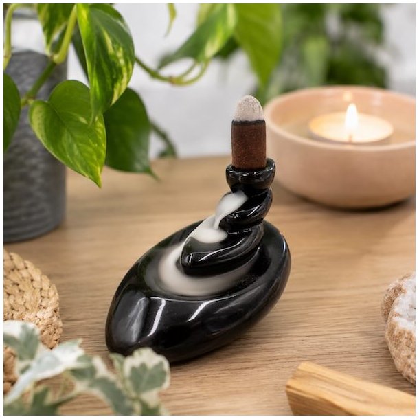 Backflow r�gelsesholder In Balance i sort glaseret keramik med vandfaldseffekt p� lyst tr�bord omgivet af gr�nne planter og stearinlys.