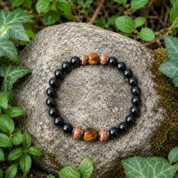 Elastisk unisex armb�nd med sort onyx, gyldent tiger�je, h�matit og tr� placeret p� en natursten omgivet af gr�nne planter